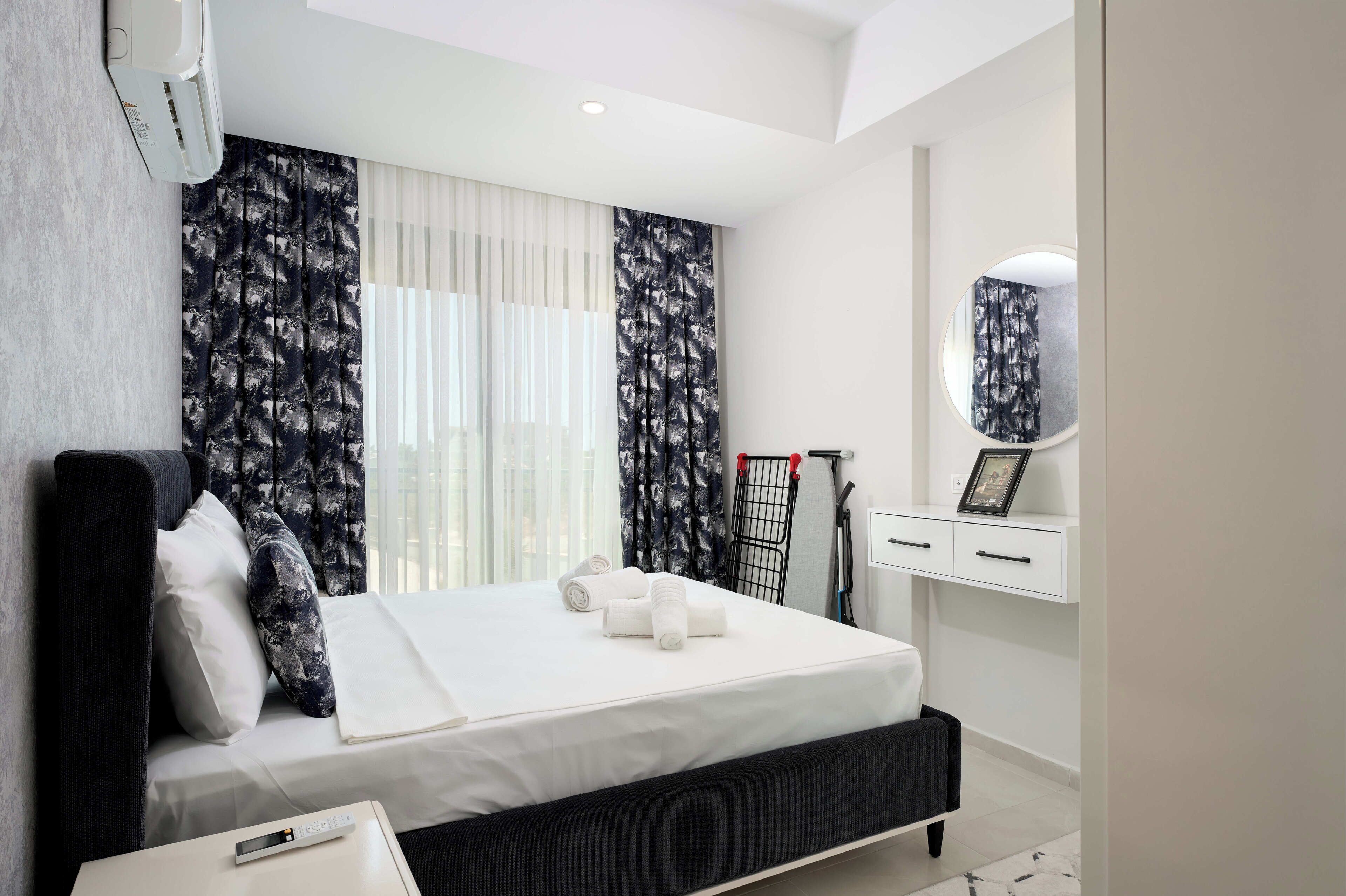 A modern hotel room with a bed, a bedside table, a mirror, and a window with curtains.