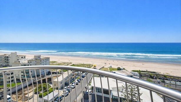 A coastal view from a balcony overlooking a beach, a parking area, and modern buildings. - Spacious Ocean View Unit — Pool, Tennis & Beaches (Surfers Paradise)