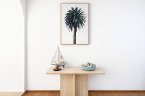 Une œuvre d’art représentant un palmier, encadrée et accrochée au mur, un petit modèle de voilier posé sur une table en bois, et un bol rempli de petits cailloux.
