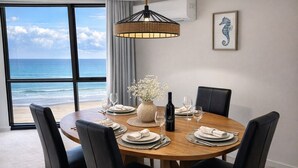 A round dining table with six chairs, a vase with flowers, wine glasses, and a bottle of wine. - Spacious Ocean View Unit — Pool, Tennis & Beaches (Surfers Paradise)