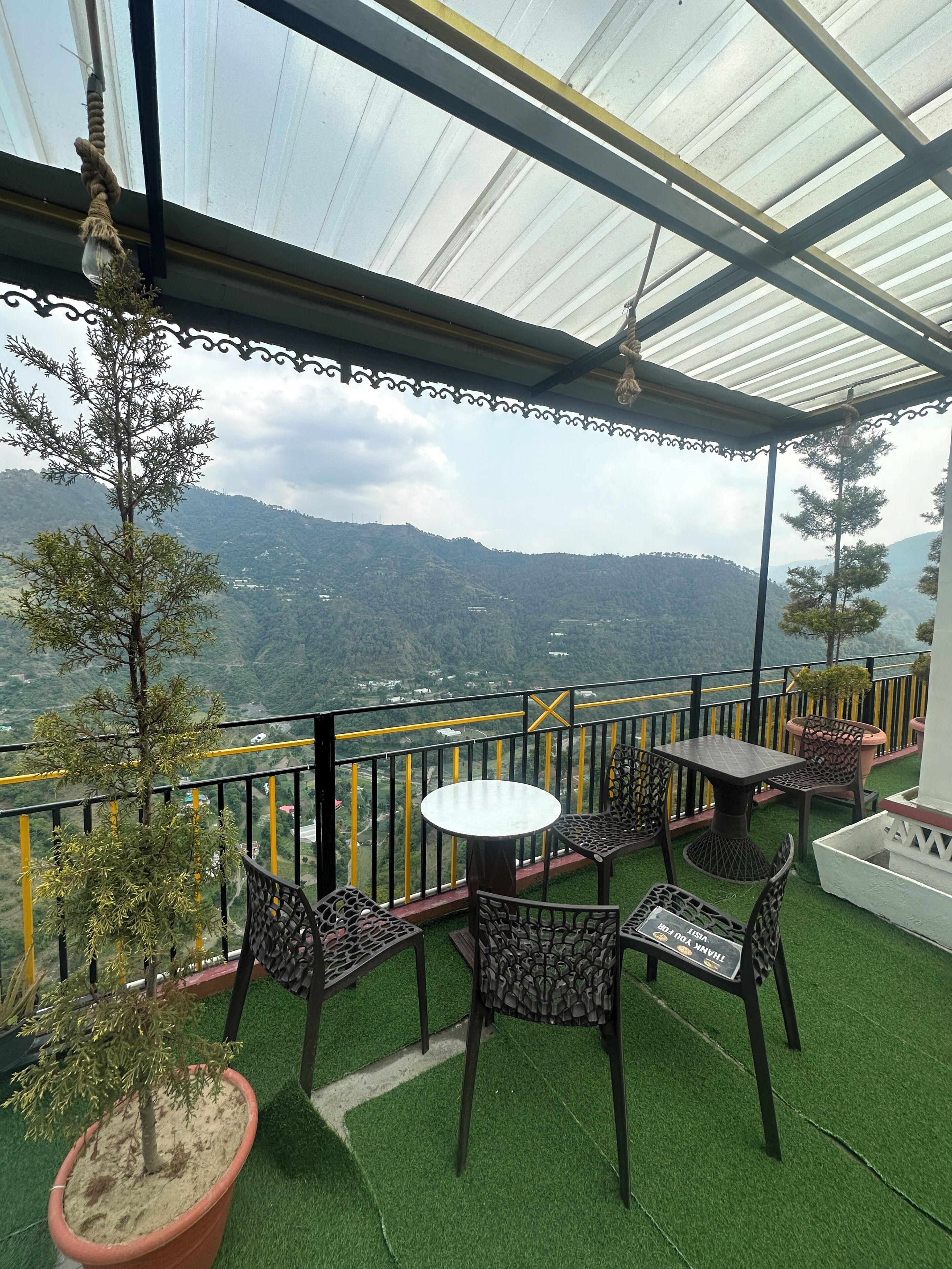 A terrace with a table and chairs, a view of a mountainous landscape, and a railing with yellow and black colors.