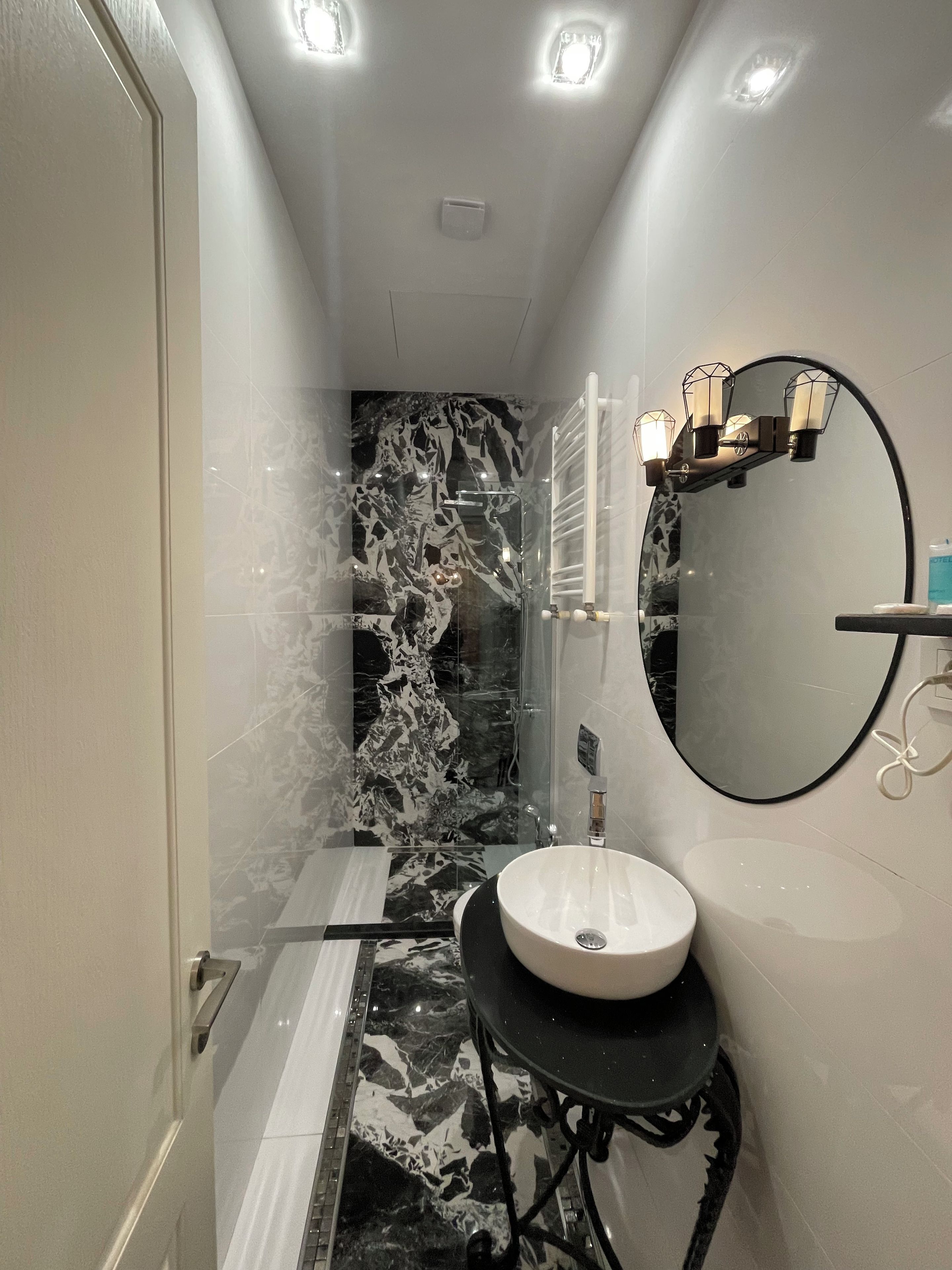 A modern bathroom with a marble shower wall, a round mirror, and a sink on a black pedestal.