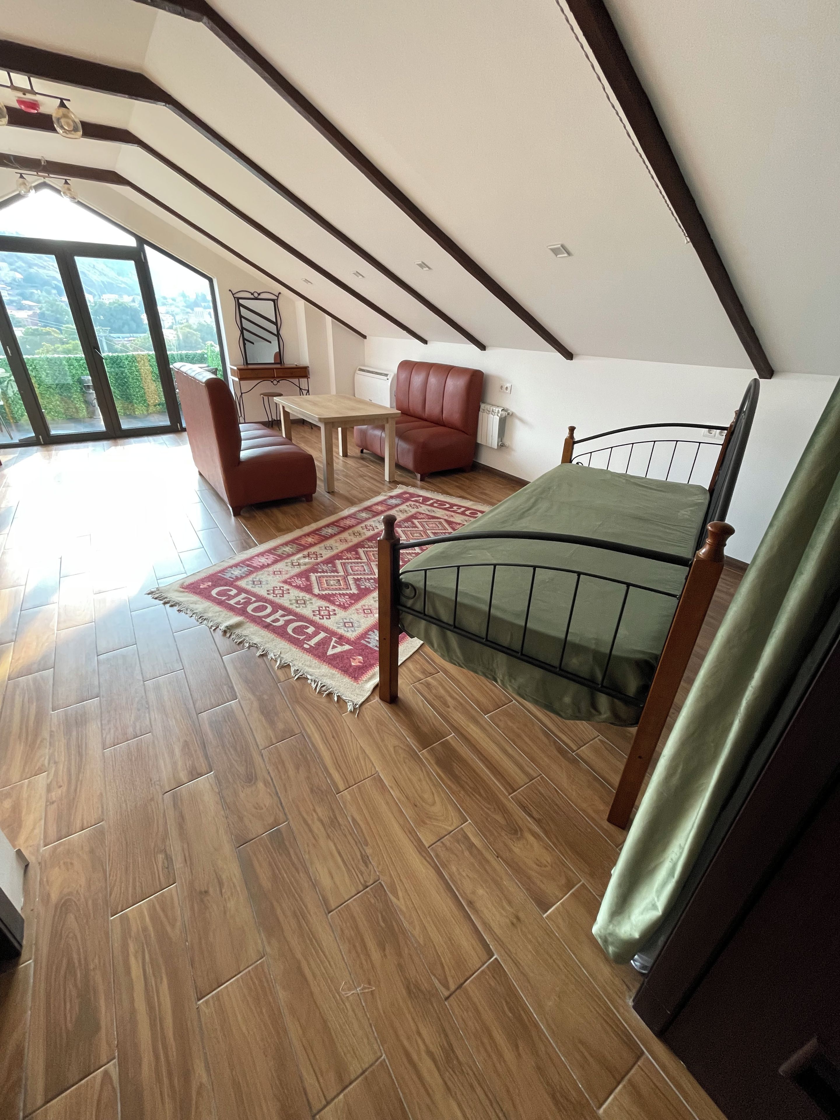 Un espacioso dormitorio en el ático con un ventanal grande, una cama, una silla, una mesa y un radiador.