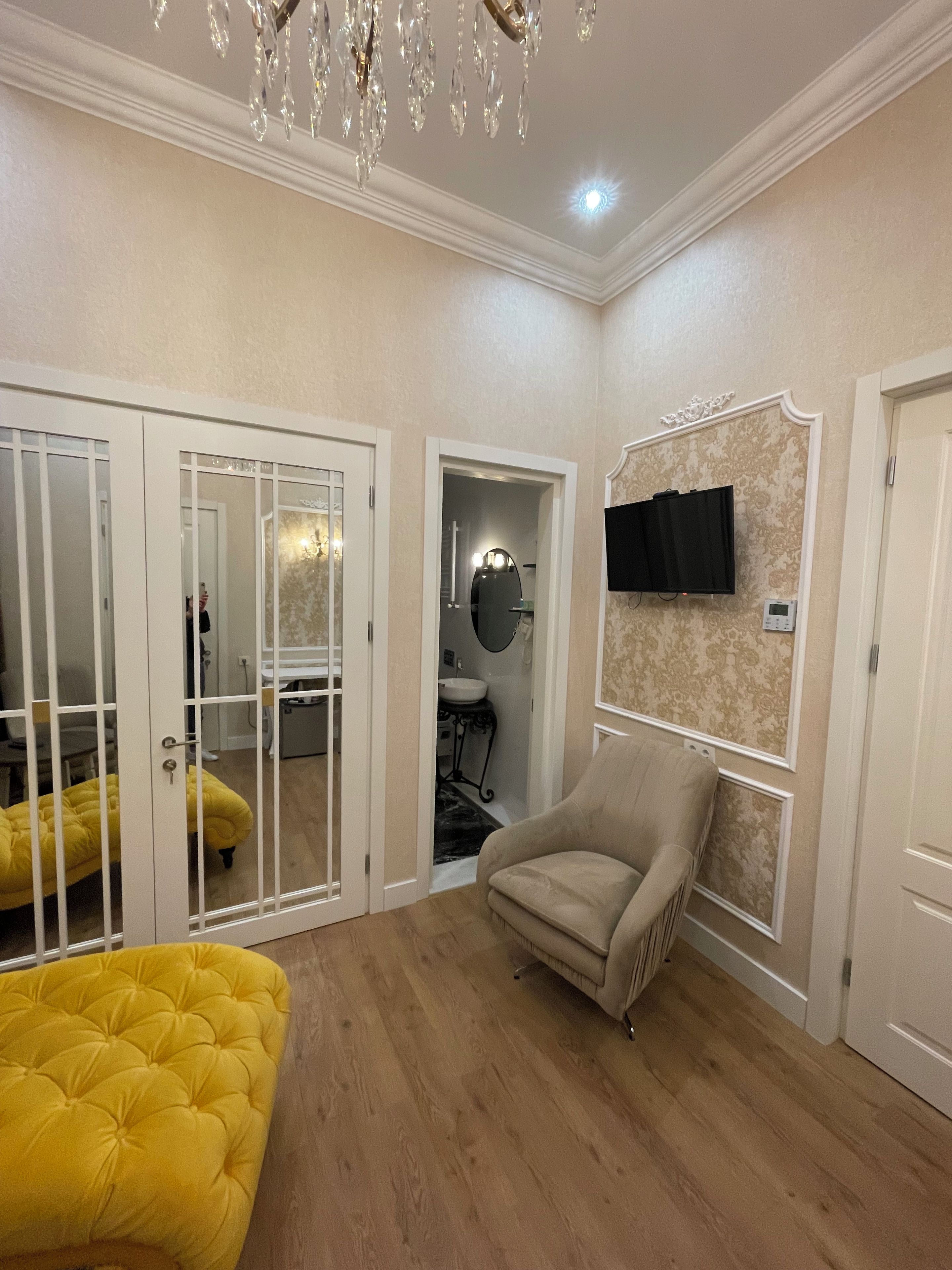 A room with a yellow tufted sofa, a beige armchair, a television mounted on the wall, and a chandelier.