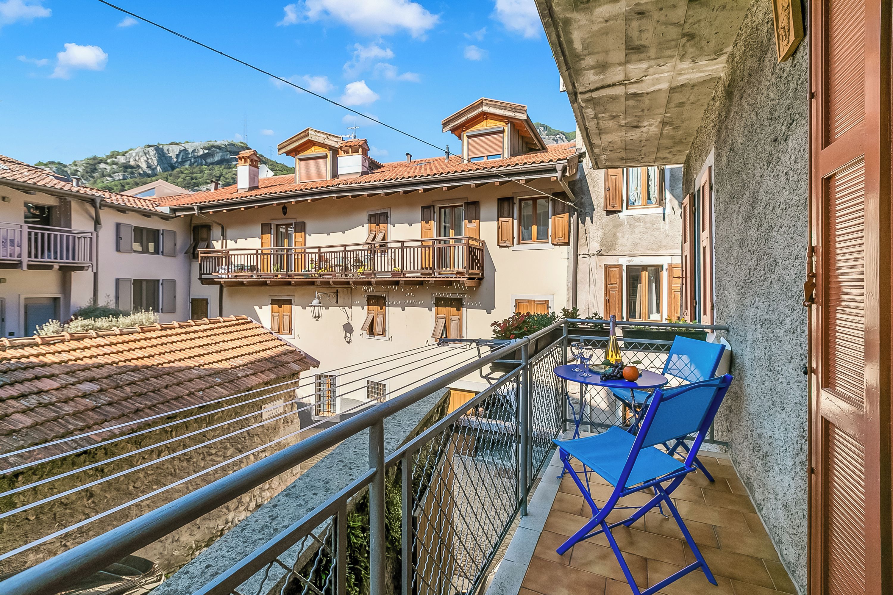 A balcony with blue chairs and a table, a stone wall, and a building with a tiled roof.