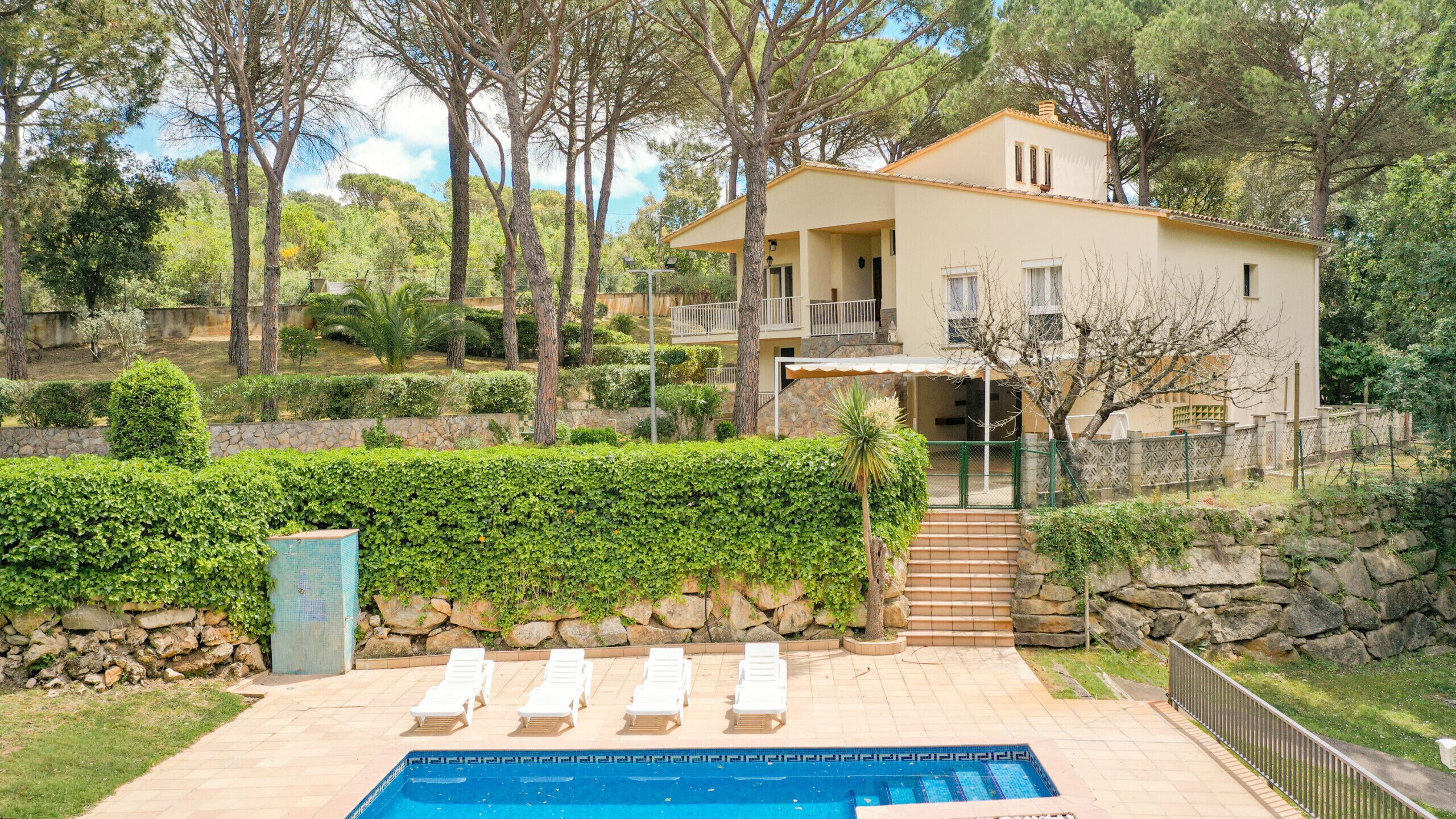 A two-story house with a swimming pool, surrounded by trees and a stone wall.