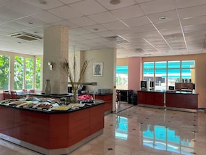 Un hall de hotel con un mostrador central tipo isla, ventanas amplias y piso de baldosas.