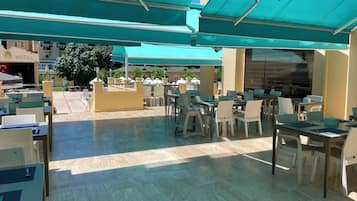 An outdoor seating area with tables and chairs, covered by blue umbrellas, and a hotel sign in the background.