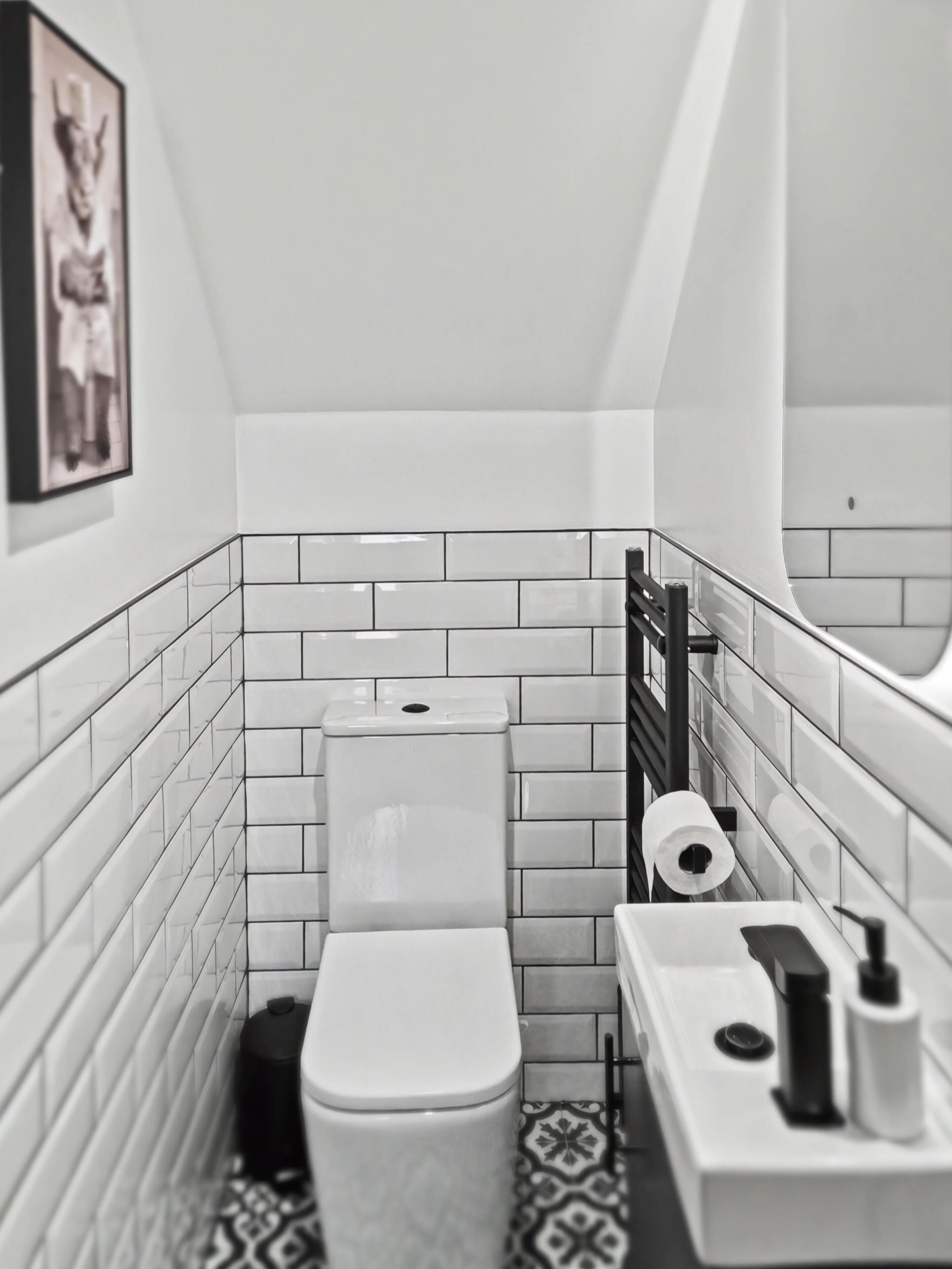 Une petite salle de bain moderne, avec des carreaux blancs, des toilettes, un lavabo avec un miroir et un sol à motifs.
