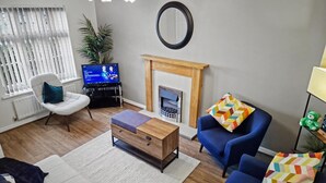 A living room with a TV, a fireplace, a blue sofa, a white armchair, and a wooden coffee table. - Stylish 2BR Solihull Stay | Near NEC | Parking | Garden (Solihull)