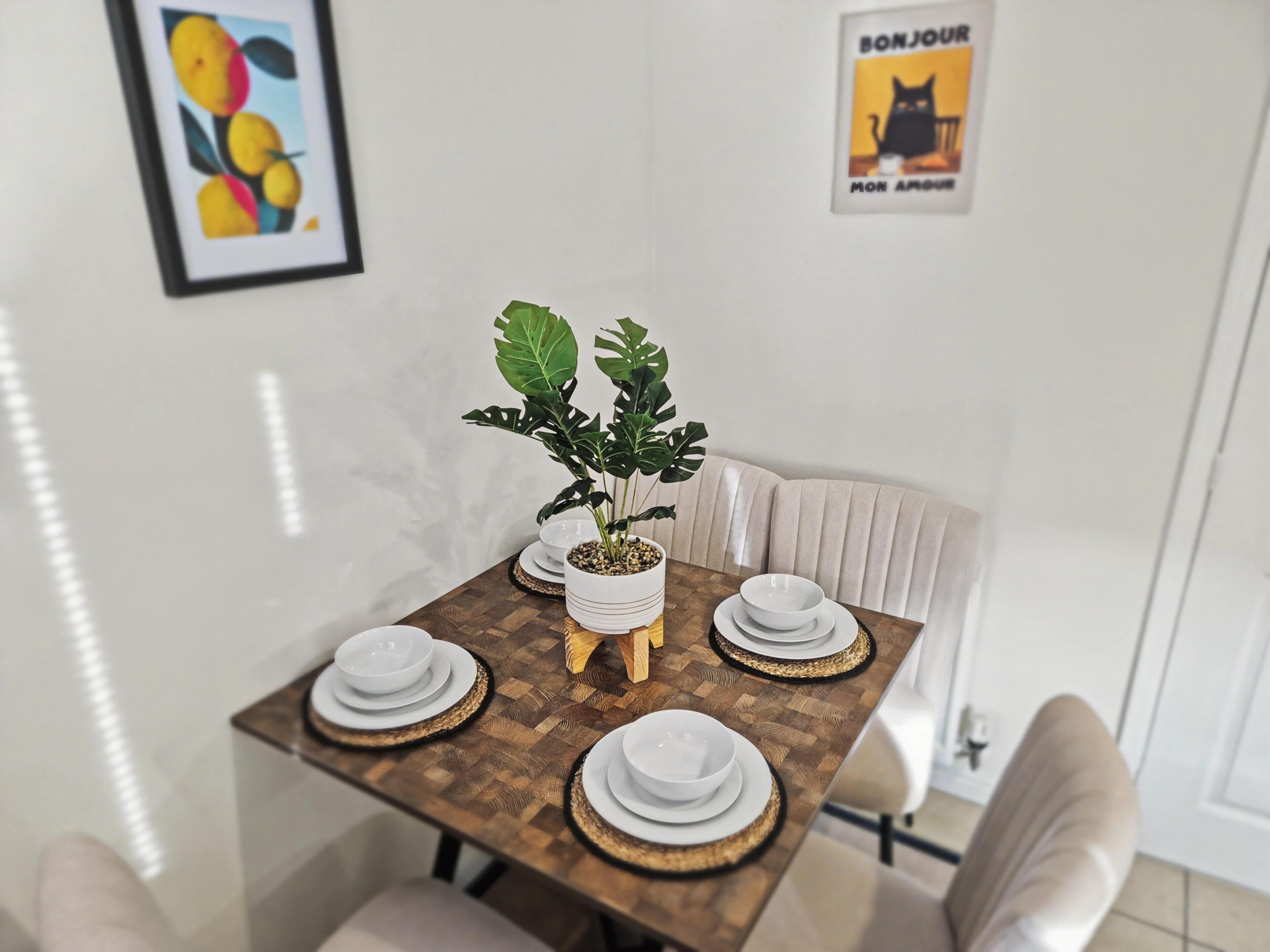 Ein Esstisch mit weißen Tellern, einem Blumentopf, einem gerahmten Kunstwerk und einem 'BONJOUR'-Schild an der Wand.