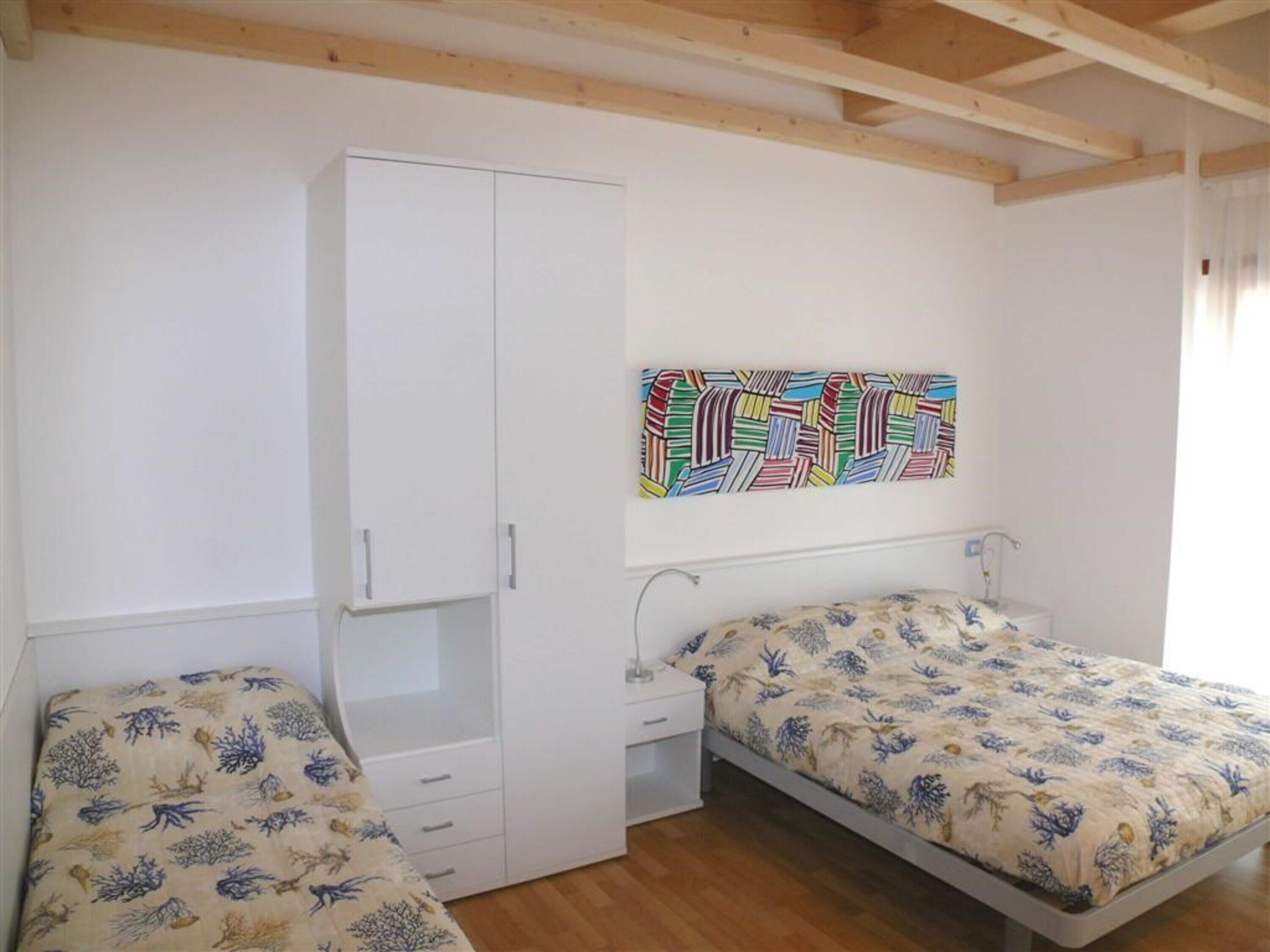 A bedroom with two beds, a wardrobe, a nightstand, and a colorful abstract painting on the wall.
