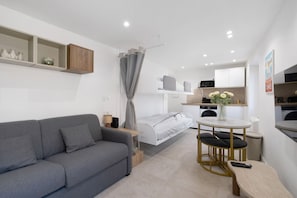 En kompakt opholdsstue med sofa, seng, lille spisebord med stole, et tekøkken og indbyggede hylder.