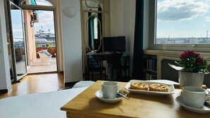 A room with a wooden table, a TV, a window with a city view, a balcony, and a dining area with pastries. - Terrazza Urania (Rome)