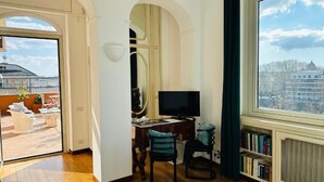 A room with a desk, chair, television, bookshelf, and a large window with a view of buildings. - Terrazza Urania (Rome)