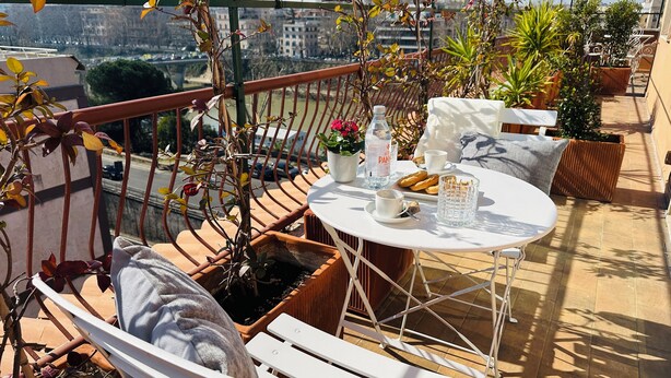 A balcony with a table set for two, a potted plant, and a view of a bridge. - Terrazza Urania (Rome)
