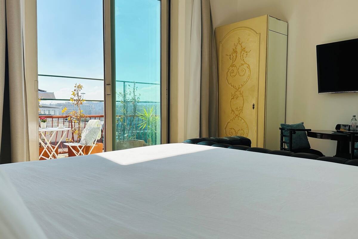A hotel room with a large bed, a desk with a chair, a TV mounted on the wall, and a balcony with outdoor furniture and plants.