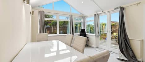 A room with a large white table, two chairs, a black bag, and a view of a garden through large windows.