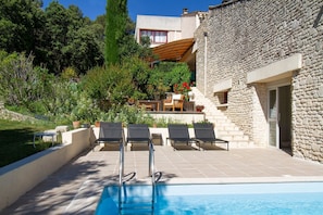Un espace piscine avec des chaises longues, un bâtiment en pierre et un jardin agrémenté de différentes plantes.
