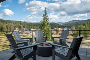 Eine Terrasse mit drei Adirondack-Stühlen und einer Feuerstelle, mit Blick auf eine Berglandschaft unter blauem Himmel.