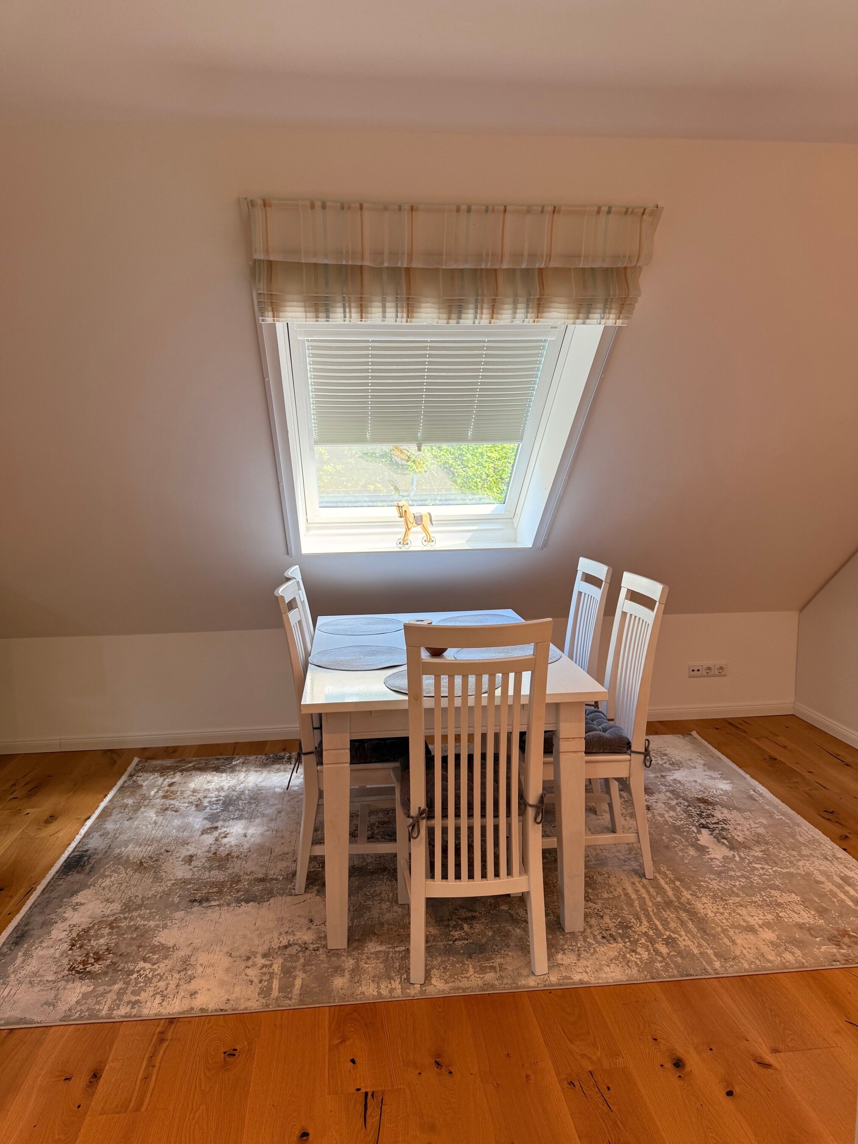 A dining area with a table and chairs, a window with blinds, and a view of greenery outside.