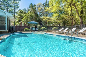 Ein Swimmingpool mit Liegestühlen, einer überdachten Sitzgruppe und einem von Zäunen umgebenen Garten mit Bäumen.