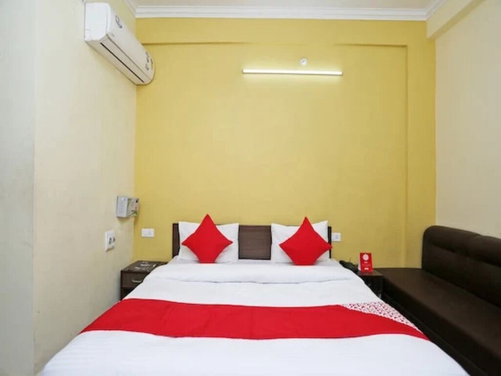 Habitación de hotel con cama, mesita de noche, colcha roja y blanca, sofá marrón y aire acondicionado instalado en la pared.