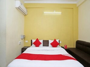 A hotel room with a bed, a nightstand, a red and white bedspread, a brown sofa, and a wall-mounted air conditioner. - Madhav Bhawan (Ayodhya)
