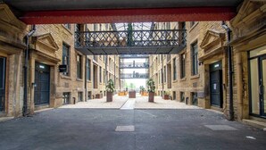 An open courtyard with a covered walkway, surrounded by brick buildings with 'WAREHOUSE' signs, and potted plants. - Charming 2-bedroom Apartment in Bradford With City Views (Bradford)