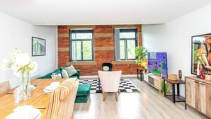 A modern living room with a wooden accent wall, a green sofa, a dining table with white plates, and a television. - Charming 2-bedroom Apartment in Bradford With City Views (Bradford)