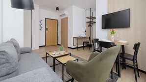 A modern living room with a grey sofa, a green armchair, a coffee table with a plant, a dining area with a table and chairs, and a mounted TV. - A comfortable studio near PGE Narodowy | A place to work (Warszawa)