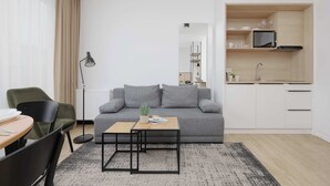A modern living room with a grey sofa, a small wooden table, a floor lamp, and a kitchen area with white cabinets and a microwave. - A comfortable studio near PGE Narodowy | A place to work (Warszawa)