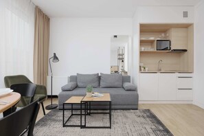 A modern living room with a grey sofa, a small wooden table, a floor lamp, and a kitchen area with white cabinets and a microwave. - A comfortable studio near PGE Narodowy | A place to work (Warszawa)