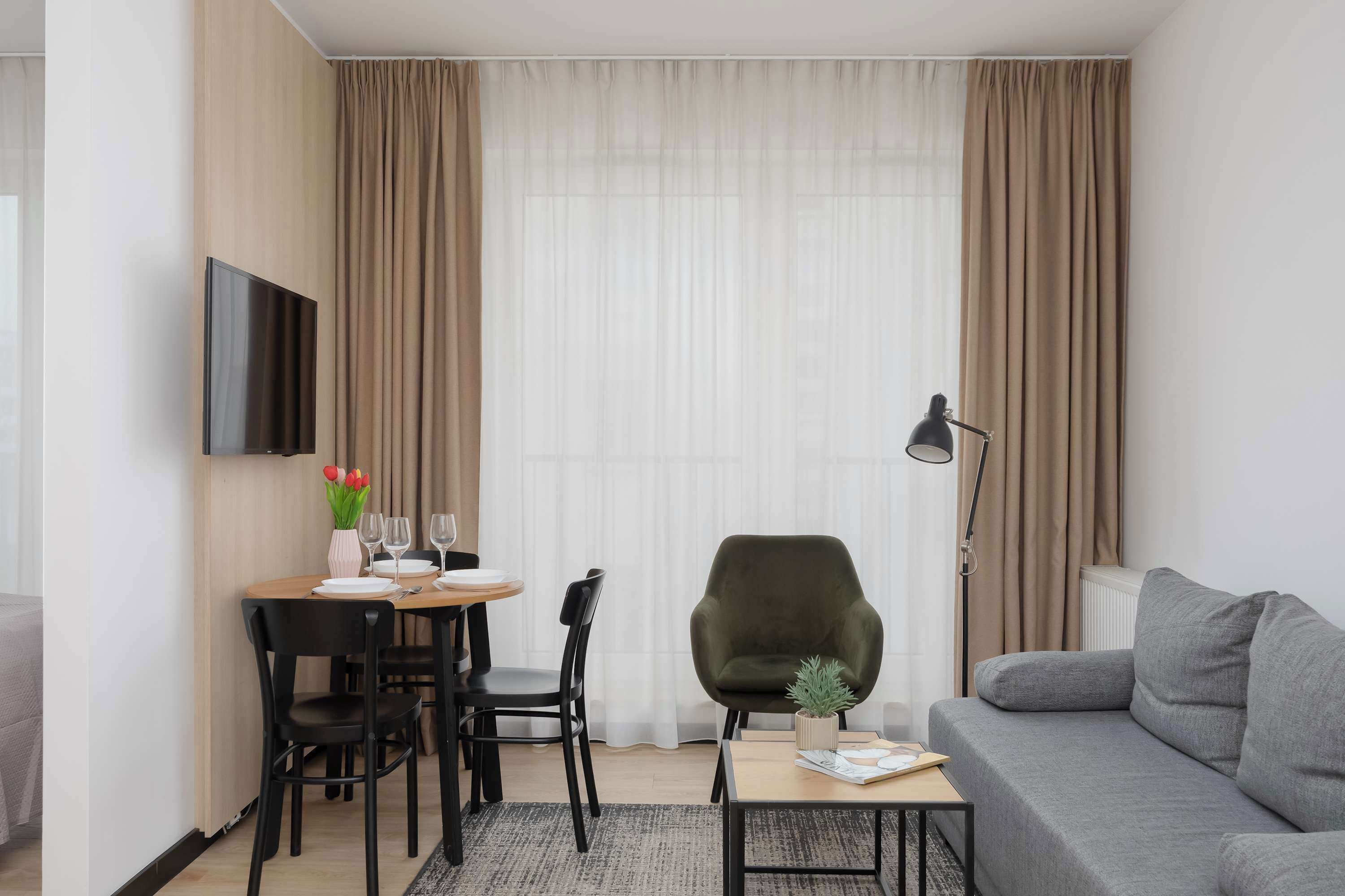 Uma sala moderna com um sofá cinza, uma mesa de jantar com cadeiras, uma televisão de tela plana na parede e uma grande janela com cortinas leves.