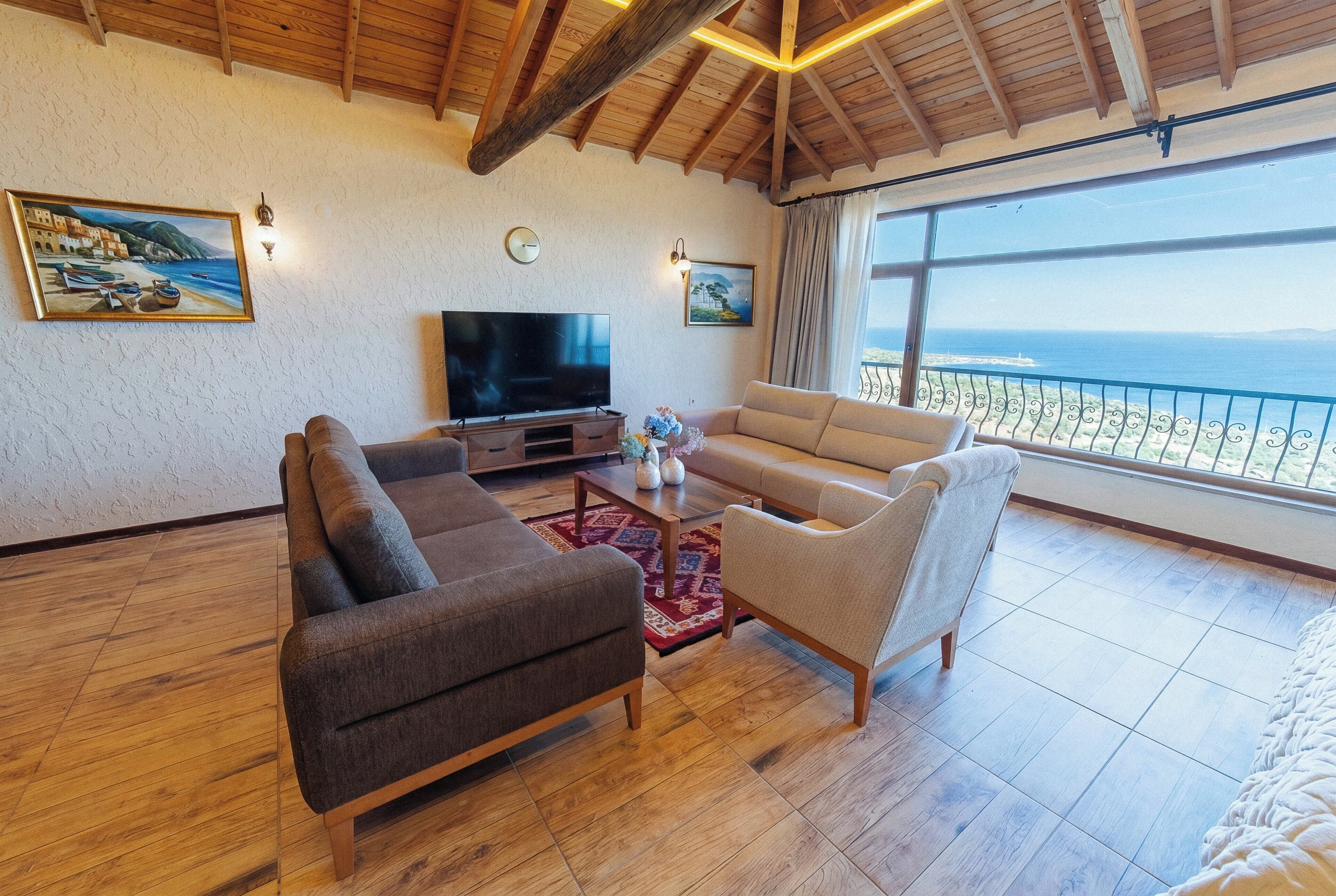 A living room with a TV, sofas, a coffee table, and a large window with a view of the sea.
