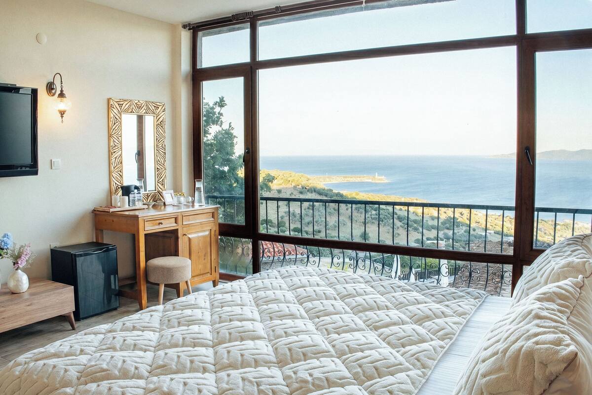 A bedroom with a large bed, a wooden desk, a mirror, and a view of the sea.