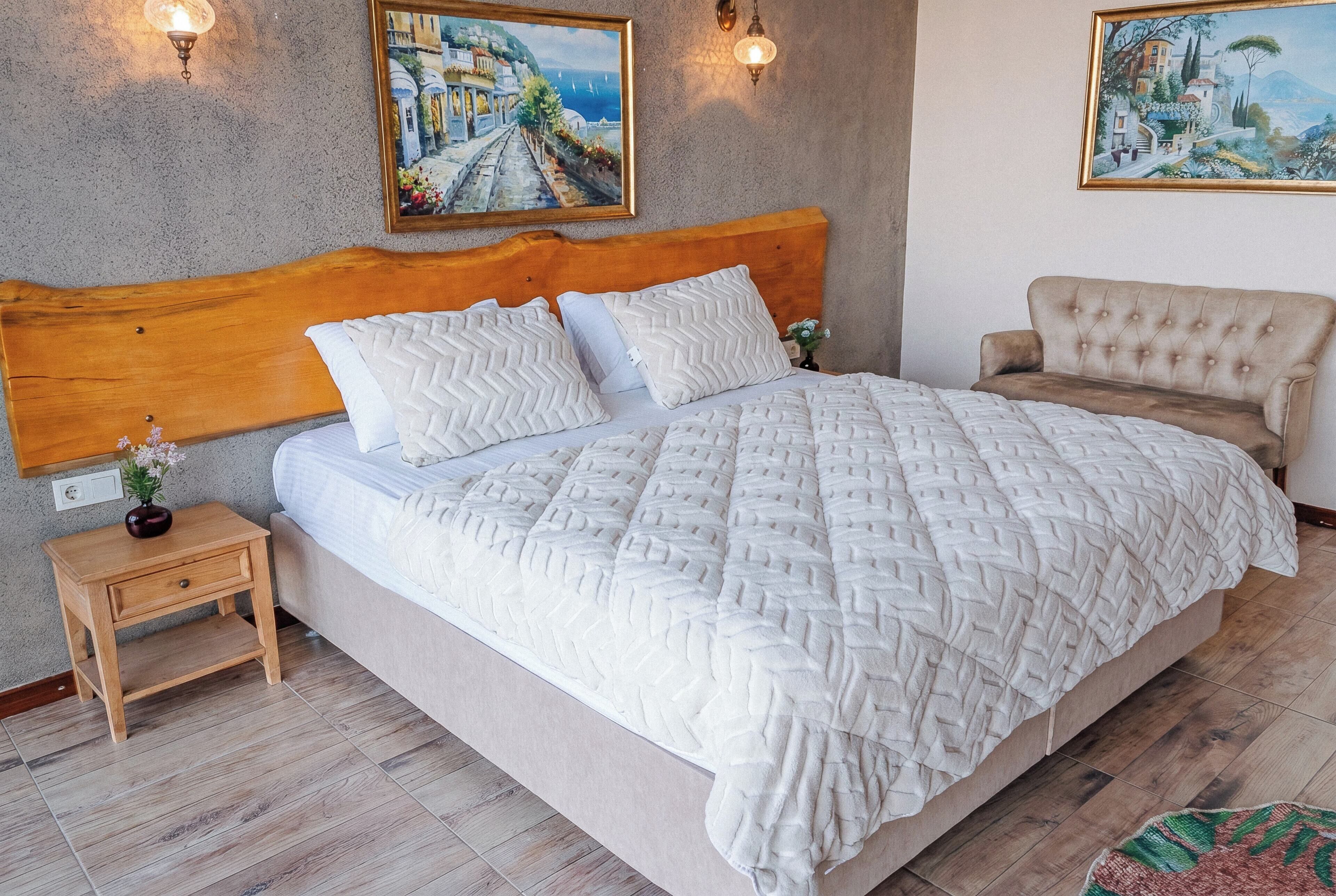 Un dormitorio con cabecera de madera, una cama grande con edredón blanco texturizado, dos lámparas fijadas a la pared y dos cuadros enmarcados.