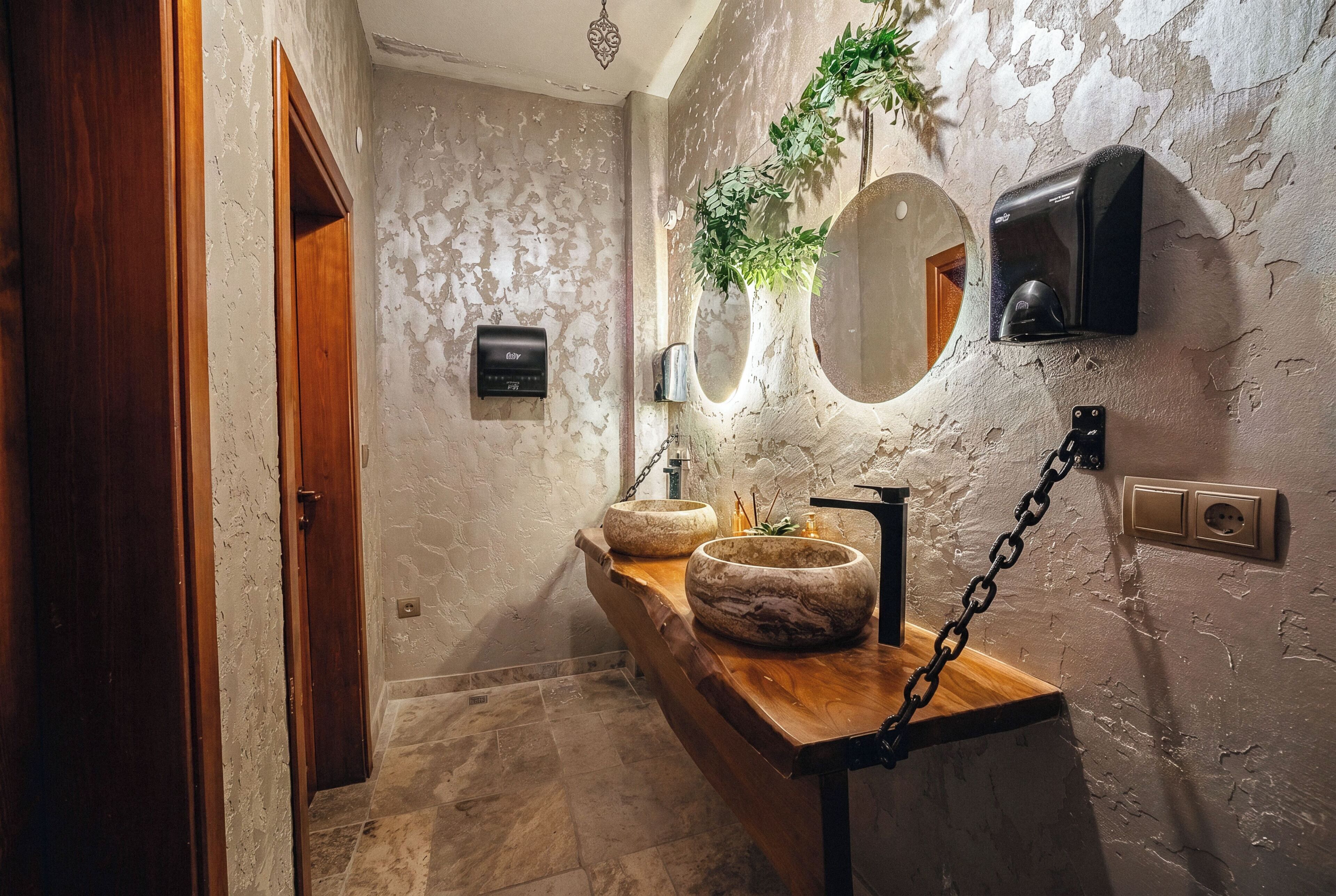 Une salle de bain avec un meuble-lavabo en bois, deux vasques en pierre, un miroir, un luminaire fixé au mur et une plante.