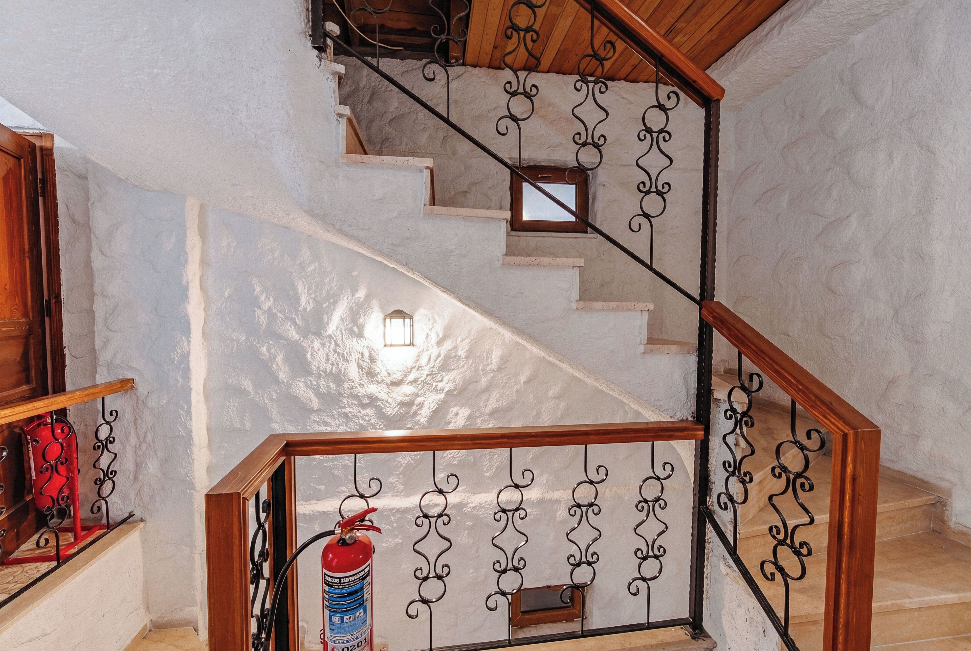 A staircase with a wooden handrail and black metal balusters, a fire extinguisher, and a small window at the top.