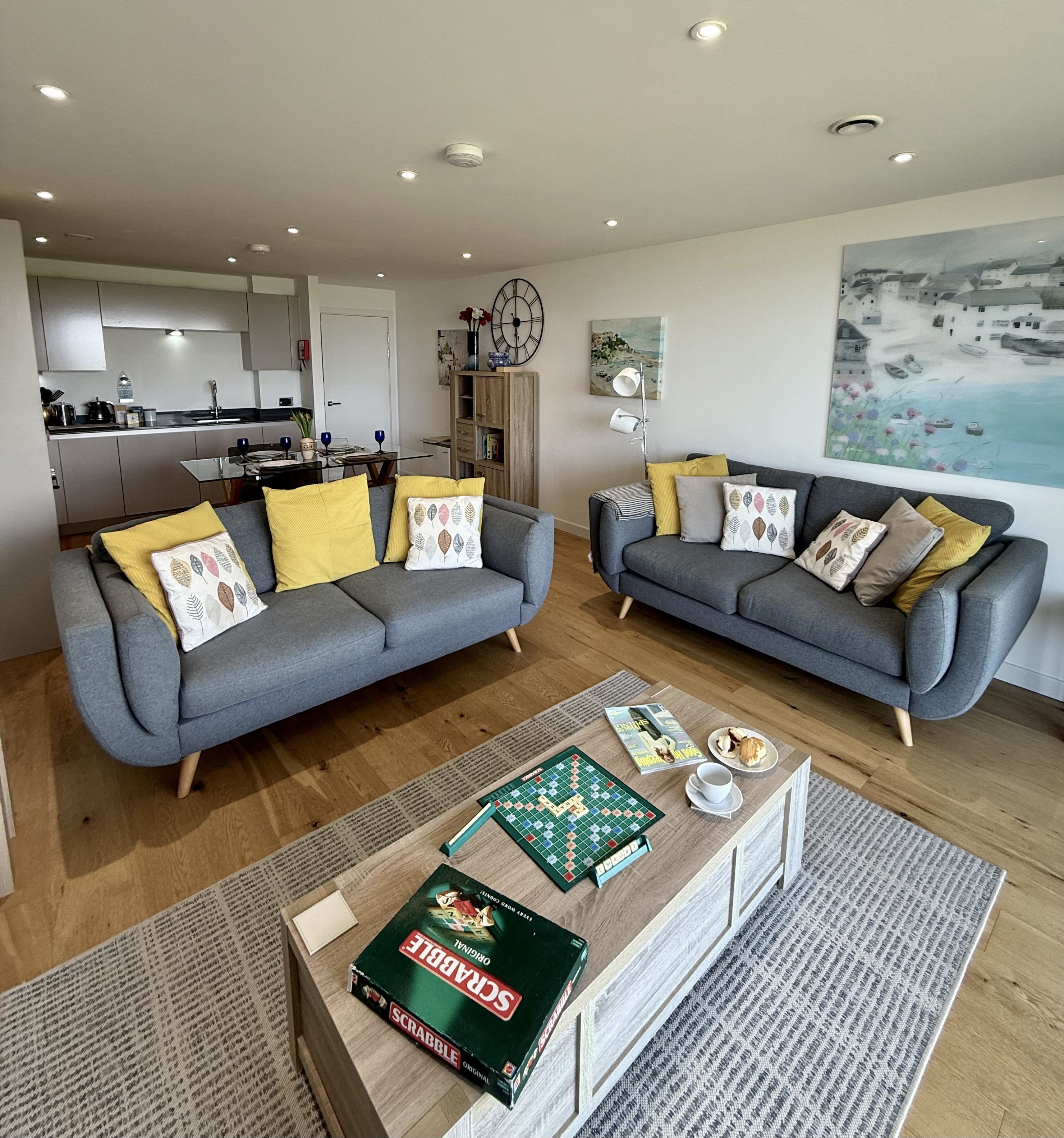 A modern living room with two grey sofas, a coffee table with a Scrabble game, and a view of a kitchen and dining area.