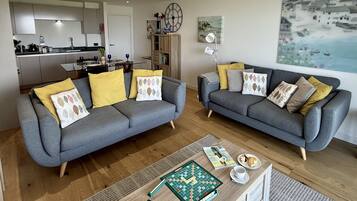A modern living room with two grey sofas, a coffee table with a Scrabble game, and a view of a kitchen and dining area.
