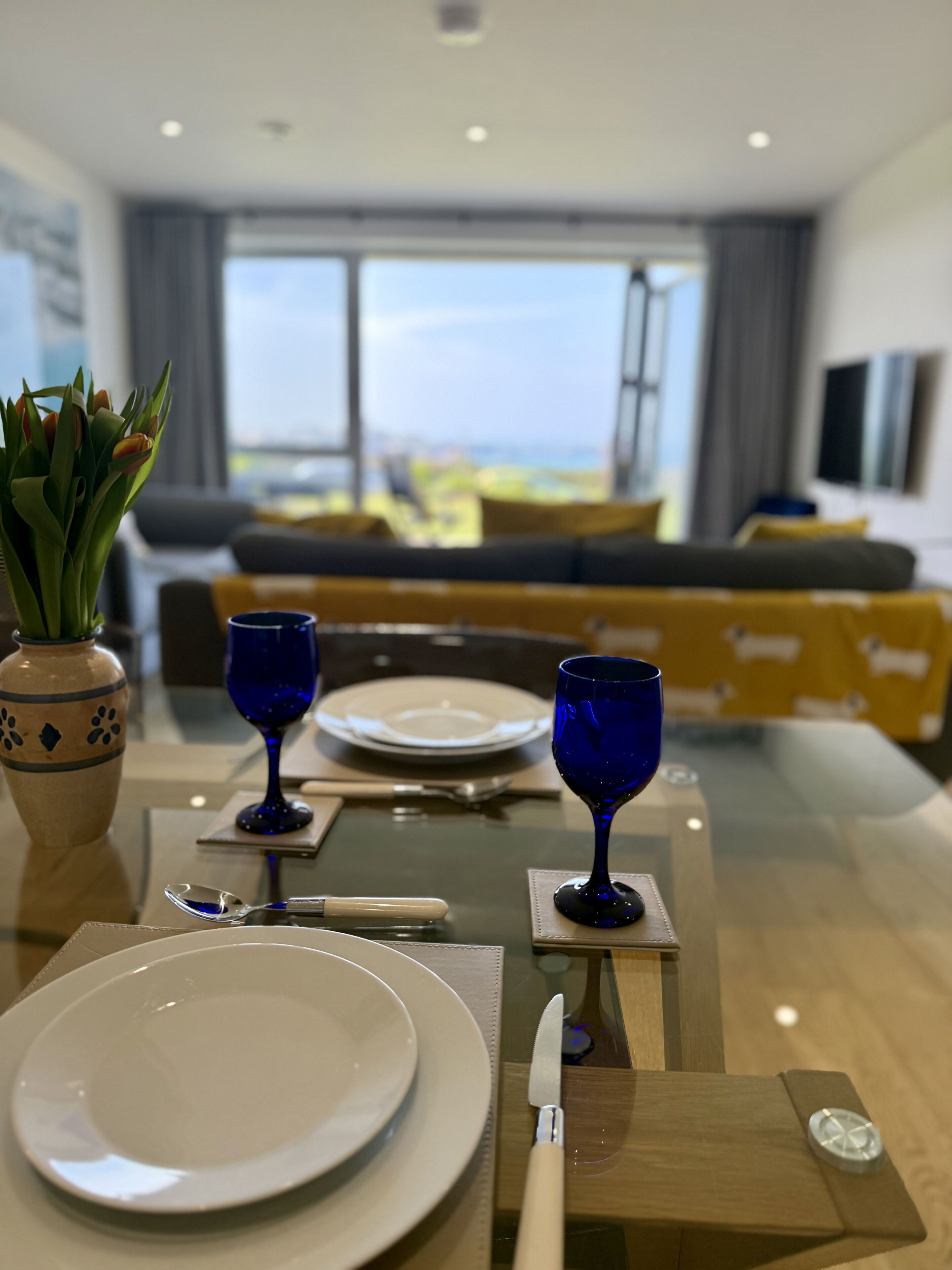 A dining table set with plates, cutlery, and blue glassware, with a view of a modern living area in the background.
