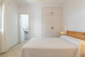 Ein Schlafzimmer mit Bett, Kleiderschrank, Nachttisch mit Lampe und angrenzendem Badezimmer, das durch eine geöffnete Tür sichtbar ist.