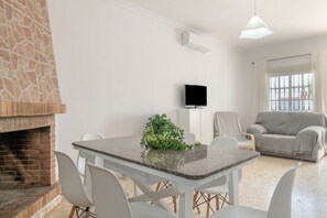 Ein Wohnzimmer mit Kamin aus Stein, einem Esstisch mit Stühlen, einem grauen Sofa und einem Fernseher an der Wand.