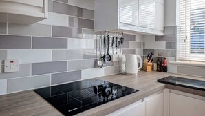 A modern kitchen with grey tiles, a black induction cooktop, and hanging utensils. - Comfortable Cottage with Decking in Castle Cary (Castle Cary)