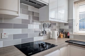 A modern kitchen with grey tiles, a black induction cooktop, and hanging utensils. - Comfortable Cottage with Decking in Castle Cary (Castle Cary)