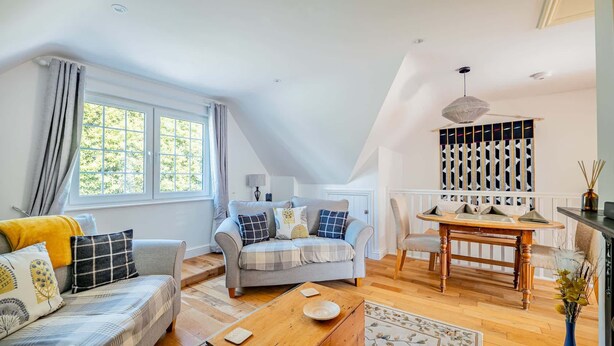 A loft-style living room with a high ceiling, a dining area with a wooden table, and a large window with curtains. - Comfortable Cottage with Decking in Castle Cary (Castle Cary)