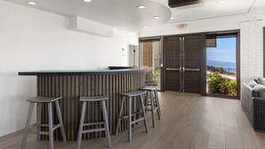 Un moderno bar con una barra de madera y cuatro taburetes, vista al mar a través de amplias puertas de cristal, y un letrero de salida sobre las puertas.