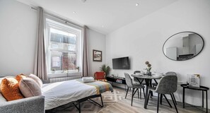 Ein modernes Wohnzimmer mit Bett, Esstisch, Stühlen, Fernseher und einem runden Spiegel.