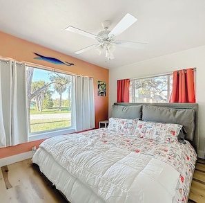 Un dormitorio con cama, ventilador de techo, ventana con cortinas y vista a palmeras.